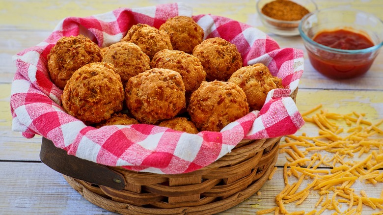a basket of hush puppies with a red gingham towel on a wooden table with dipping sauces.
